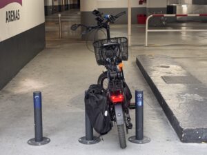 Entrée du parking Indigo Arénas Aéroport : un cycliste avec une sacoche arrière arrive à peine à franchir l'accès protégé par des bornes de sécurité très serrées. Ce passage étroit limite l'ergonomie et rend l'accès difficile aux vélos équipés ou aux modèles cargos, malgré une implantation en rez-de-chaussée.