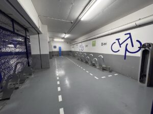 Intérieur du local vélo sécurisé Indigo Arénas Aéroport. L'espace comprend des râteliers métalliques gris pour attacher les vélos par le cadre et la roue. On aperçoit également des casiers de rangement numérotés pour l'équipement des cyclistes.