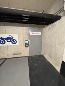 Entrée sécurisée du parking vélo Indigo Joia Méridia. Une large porte métallique, signalée par la mention Cyclopark, permet d'accéder au local depuis l'intérieur du parking souterrain, à côté du stationnement réservé aux motos. L'accès est contrôlé par un lecteur de badge situé sur le montant de la porte pour garantir la sécurité.