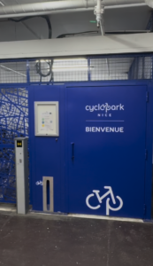 Entrée du local vélo sécurisé Saleya (Indigo) à Nice. Une porte métallique bleue avec un grand logo de vélo et la mention « Cyclopark » s'ouvre sur un espace propre et bien éclairé. Un boîtier de contrôle d'accès est visible sur la grille à gauche pour une ouverture par badge.