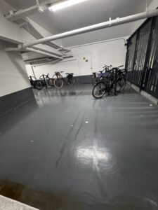 Vue intérieure du parking vélo sécurisé Promenade des Arts à Nice. Le local est clôturé avec une grille grise, propre et bien éclairé, équipé d'arceaux métalliques gris fixés au sol, permettant d'attacher les vélos par le cadre et la roue pour une sécurité optimale. Des vélos de ville avec paniers et sacoches sont stationnés et accrochés aux arceaux. Le sol est en béton gris et les murs blancs, offrant un espace de stationnement dégagé et moderne.