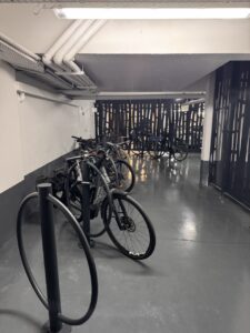 Vue intérieure du parking vélo sécurisé Promenade des Arts à Nice. Le local est clôturé avec une grille grise, propre et bien éclairé, équipé d'arceaux métalliques gris fixés au sol, permettant d'attacher les vélos par le cadre et la roue pour une sécurité optimale. Des vélos de ville avec paniers et sacoches sont stationnés et accrochés aux arceaux. Le sol est en béton gris et les murs blancs, offrant un espace dégagé et moderne où stationner son vélo en sécurité.