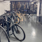 Vue intérieure du parking vélo sécurisé Promenade des Arts à Nice. Le local est clôturé avec une grille grise, propre et bien éclairé, équipé d'arceaux métalliques gris fixés au sol, permettant d'attacher les vélos par le cadre et la roue pour une sécurité optimale. Des vélos de ville avec paniers et sacoches sont stationnés et accrochés aux arceaux. Le sol est en béton gris et les murs blancs, offrant un espace dégagé et moderne où stationner son vélo en sécurité.