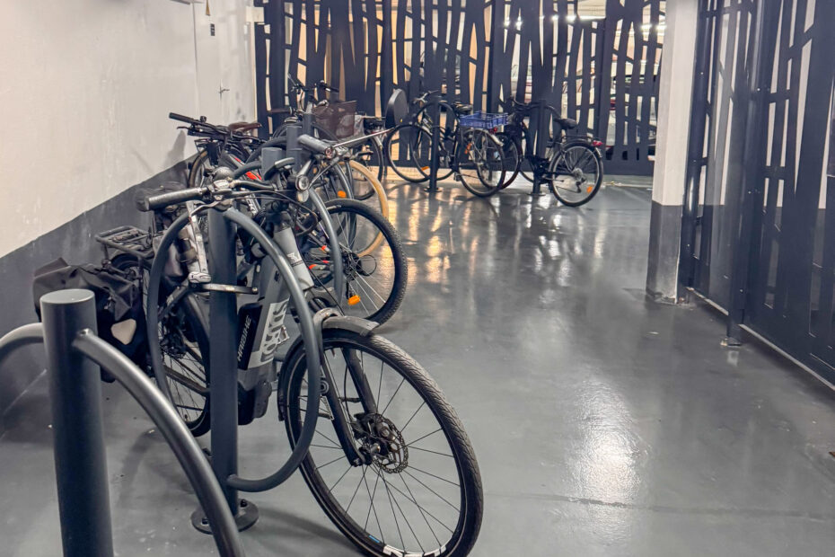 Vue intérieure du parking vélo sécurisé Promenade des Arts à Nice. Le local est clôturé avec une grille grise, propre et bien éclairé, équipé d'arceaux métalliques gris fixés au sol, permettant d'attacher les vélos par le cadre et la roue pour une sécurité optimale. Des vélos de ville avec paniers et sacoches sont stationnés et accrochés aux arceaux. Le sol est en béton gris et les murs blancs, offrant un espace dégagé et moderne où stationner son vélo en sécurité.