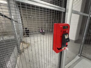 Porte d'accès sécurisée d'un local vélo Q-Park Gare du Sud. Le local est clôturé par une grille métallique, et une borne rouge de contrôle d'accès par badge est visible. À l'arrière-plan, on aperçoit des vélos attachés à des râteliers 'pince-roue', qui ne permettent pas d'attacher le cadre en toute sécurité.